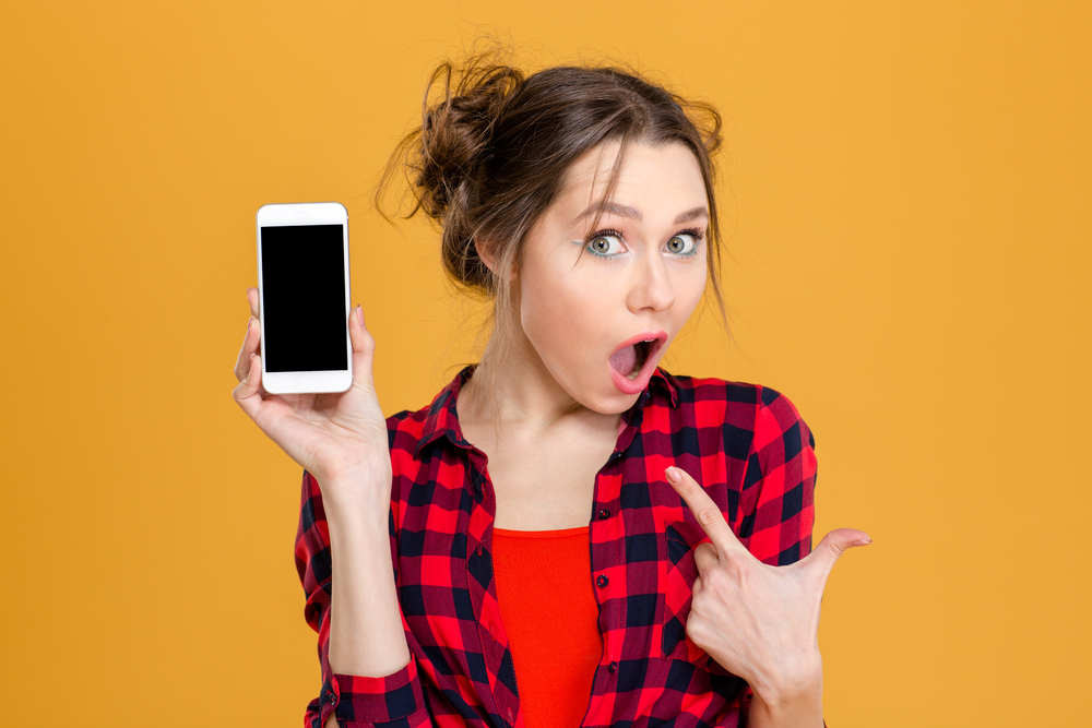 Portrait of a young amazed woman showing blank smartphone screen over yellow background. Focus on smartphone